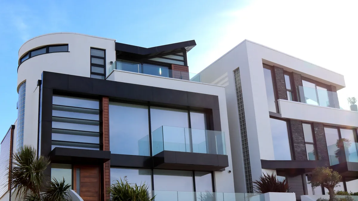 A modern, multi-story house with white walls, black trim, and large windows, featuring balconies and a flat roof, set against a blue sky wit