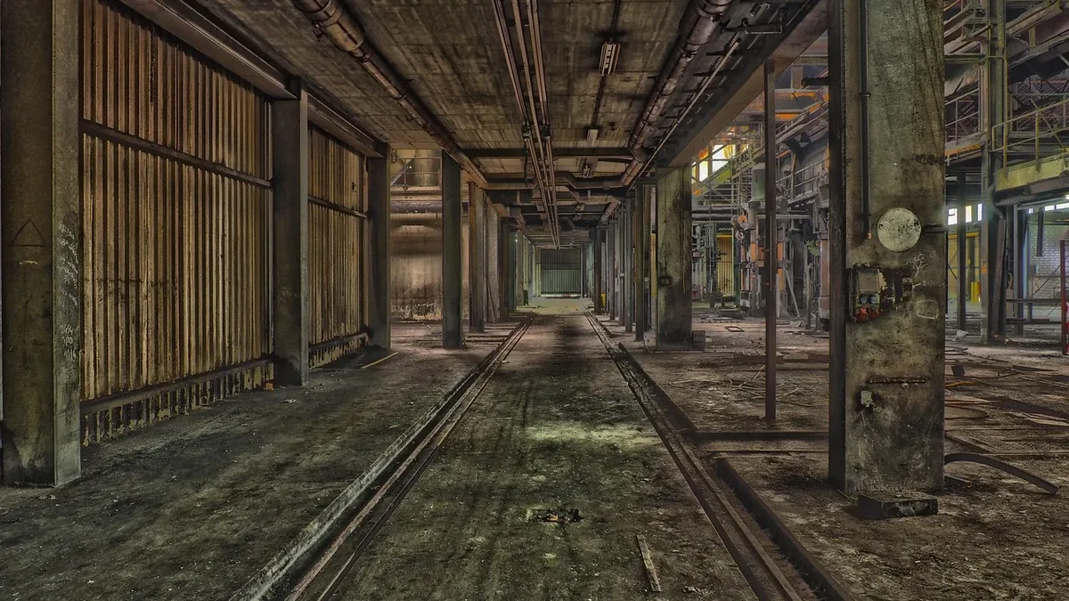 An empty, dimly lit industrial warehouse with exposed pipes and tracks on the floor.