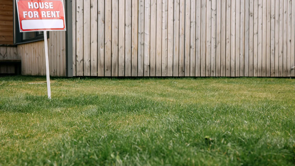 A "HOUSE FOR RENT" sign in a grassy area in front of a wooden fence.